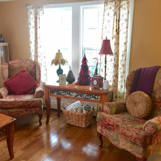 A cozy living room with armchairs, decorative pillows, and a wooden floor, featuring a table between two windows adorned with lamps and ornaments.