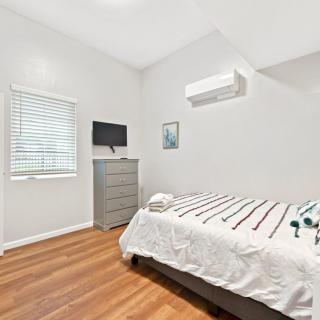 A minimalist bedroom with wooden flooring, featuring a bed, dresser, wall-mounted television, and air conditioning unit.