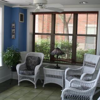 A cozy sunroom featuring wicker chairs, a matching table, and lush greenery.