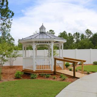 A charming garden gazebo with a wooden ramp, surrounded by greenery and bordered by a white fence on a sunny day.