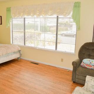 A cozy bedroom with a large window, featuring a bed and an armchair on a wooden floor.