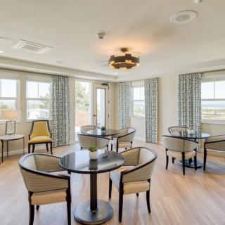 A bright and inviting dining area with round tables and upholstered chairs, featuring large windows and curtains.