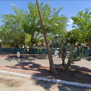 A view of a building surrounded by trees and cactus, with a parking area in the foreground.