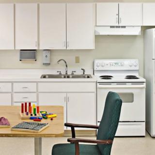 A small kitchen area with white cabinets, a stove, refrigerator, and sink. In the foreground, there's a table with toys and a chair.