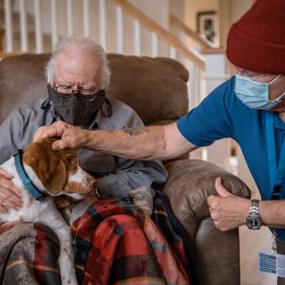 An elderly person wearing a mask sits on a couch, petting a dog with the assistance of a caregiver.
