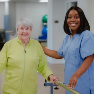 A nurse assists a senior woman during a rehabilitation session, both smiling and engaging in care.