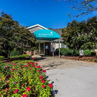 A building entrance with a blue awning surrounded by landscaped flowers and trees.