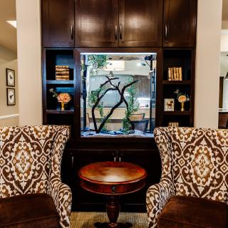 A cozy living room with patterned armchairs flanking a central table and aquarium, surrounded by dark wood bookshelves.