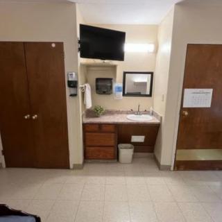 A hotel room interior showing a bathroom sink with a mirror above, a television mounted on the wall, and two doors flanking the dresser area.
