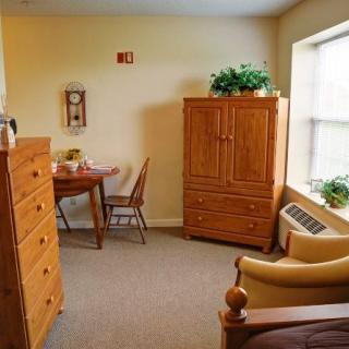 A cozy bedroom with wooden furniture, including a wardrobe, chest of drawers, table, and chairs, complemented by indoor plants and natural light from the window.
