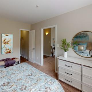 A cozy bedroom with a patterned bedspread, dresser with an oval mirror, and decorative artwork on the wall.