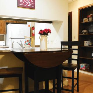 A cozy kitchen and dining area with bar stools, a small table adorned with flowers, and a bookshelf in the background.