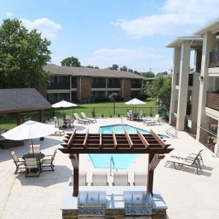 A view of an outdoor swimming pool surrounded by a patio area with sun umbrellas and apartment buildings in the background.