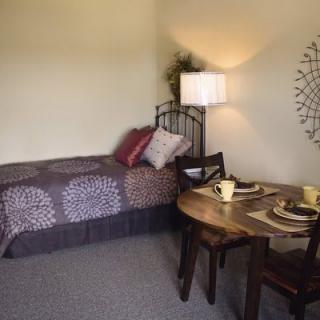A cozy bedroom with a single bed and a small dining table, complemented by warm lighting and decorative wall art.
