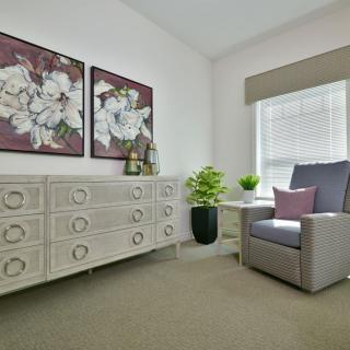 A cozy living room corner featuring a gray armchair, a large dresser with intricate patterns, floral artwork, and a green plant by the window.