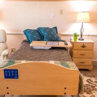 A neatly arranged hospital bedroom featuring a bed with pillows, an adjustable tray, a nightstand with a lamp, and a comfortable chair.