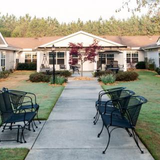 A serene outdoor pathway lined with chairs, leading to a residential building surrounded by greenery.