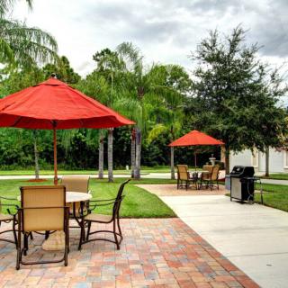 A peaceful outdoor patio area with tables, chairs, red umbrellas, a grill, and lush green surroundings.