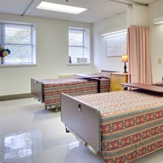 A clean and well-lit hospital patient room featuring two beds, a bedside table, and large windows.