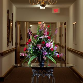 A beautifully arranged bouquet of flowers sits on a table in a dimly lit hallway, illuminated by a chandelier and accompanied by an exit sign above the door.