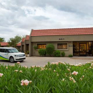 A building with a tiled roof next to a parked car, surrounded by landscaped greenery and flowers under a cloudy sky.
