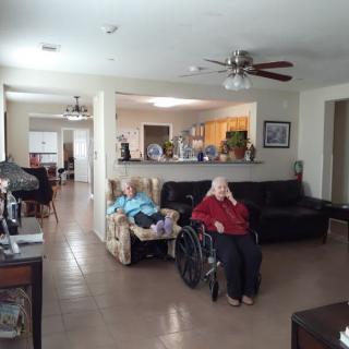 An elderly person in a wheelchair is sitting in an open-plan living room with a view into the kitchen.