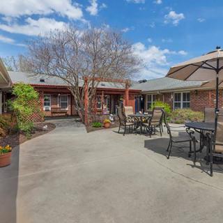 A spacious courtyard with outdoor seating, surrounded by buildings and trees under a blue sky with clouds.