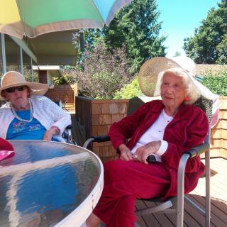 Two elderly women sitting on a patio under an umbrella, wearing sun hats.