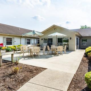 A sunny patio area with tables and chairs in front of a building, surrounded by landscaping.