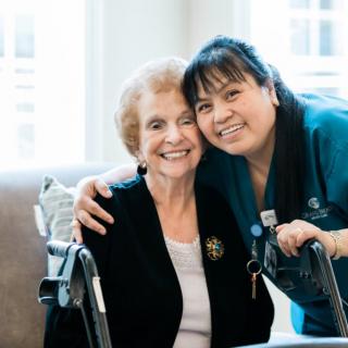 An elderly woman and her caregiver share a warm embrace, smiling together.
