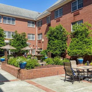 A sunny courtyard with patio furniture, surrounded by a brick building and lush greenery.