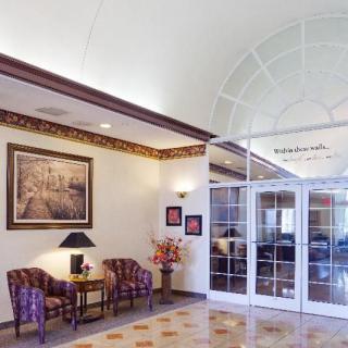 A hotel lobby featuring comfortable chairs, a small table with a lamp, decorative flowers, and framed artwork on the wall.