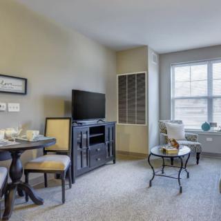 A cozy living room with a small dining table and chairs, TV stand, and a window providing natural light.
