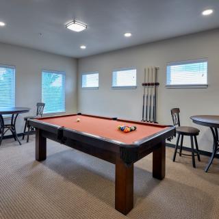 A cozy game room featuring a pool table with a set of billiard balls arranged, surrounded by bar stools and windows for natural light.