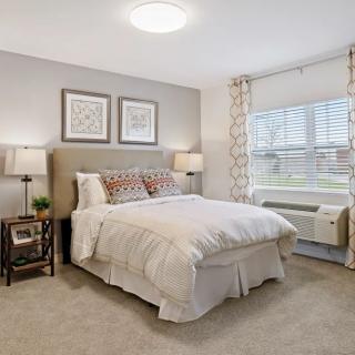A modern bedroom with neutral tones, featuring a comfortable bed, lamps, and large windows.