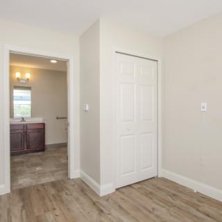 An empty bedroom with a view of a bathroom through the doorway, featuring hardwood floors and neutral-colored walls.