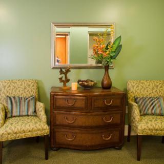 A cozy living room corner with a wooden dresser, two patterned chairs, and a decorative mirror above.