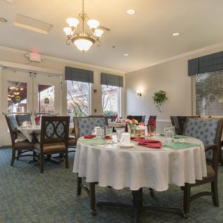 An elegant dining room with tables set for a meal, featuring a chandelier and large windows.