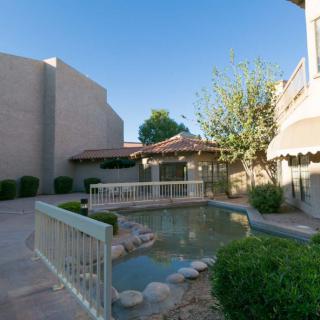 A tranquil courtyard featuring a small pond with a bridge, surrounded by greenery and buildings under a clear blue sky.