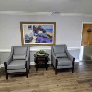 A cozy waiting area featuring two grey chairs, a small table, and decorative artwork on the wall.