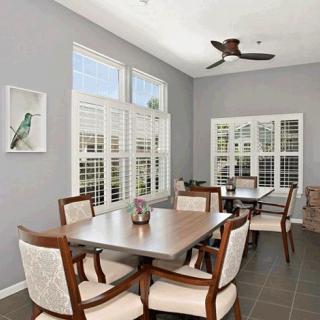 A bright dining room with a wooden table and chairs, featuring large windows with shutters and a ceiling fan.