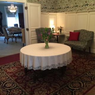 A cozy room with a round table covered in a white cloth, surrounded by upholstered chairs and decorated with flowers.