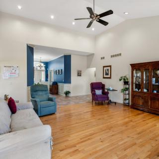 A spacious living room with wooden flooring, featuring a couch, armchairs, ceiling fan, and a display cabinet.