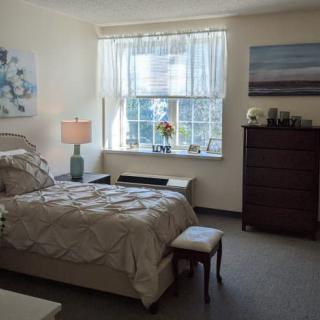 A cozy bedroom with a neatly made bed, artwork on the walls, and light streaming in through a window.