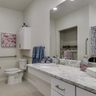A modern bathroom featuring a marble countertop with a sink, a large mirror, towels hanging on the wall, and decorative elements.