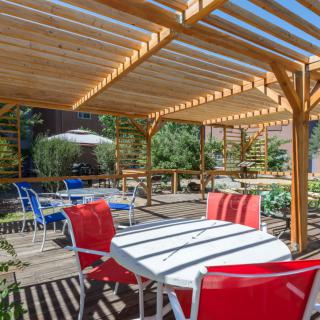 A sunny outdoor patio featuring colorful chairs under a wooden pergola, surrounded by lush greenery.