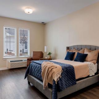 A cozy bedroom with a neatly made bed, an armchair by the window, and wooden flooring.