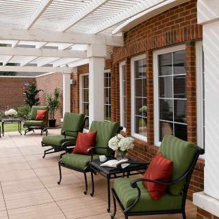 A stylish patio area with comfortable seating under a pergola, featuring green cushions and red pillows against a brick wall.