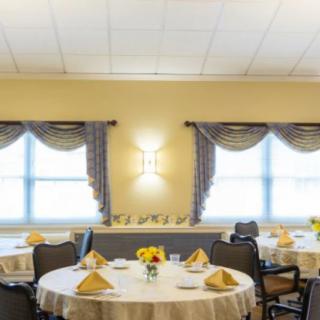A well-decorated dining room featuring round tables with neatly arranged table settings, set beside large windows with draped curtains.