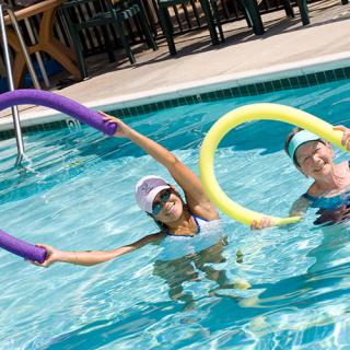 Two individuals enjoying a water exercise with pool noodles in an outdoor swimming pool.